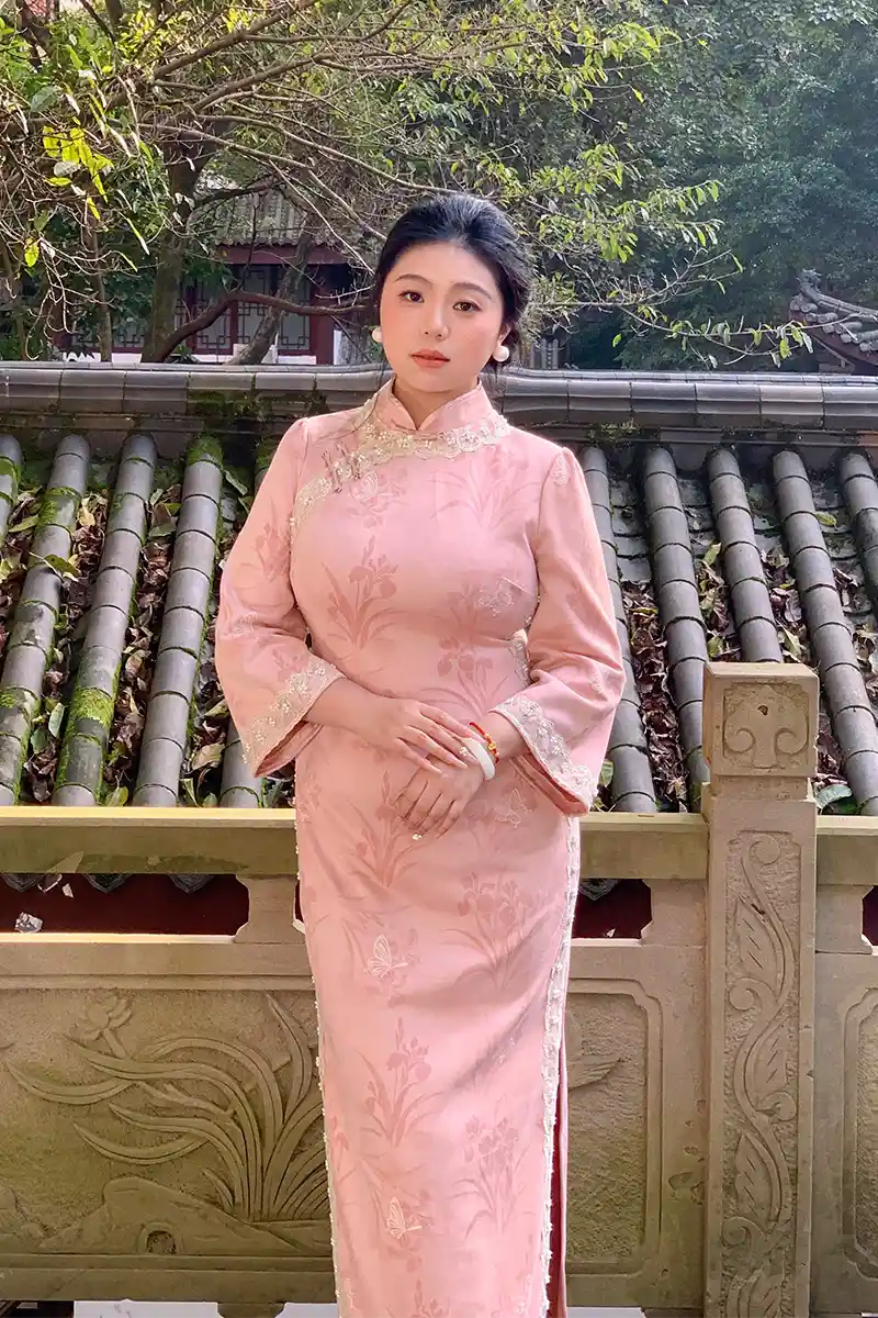 Woman in a pink traditional Plus Size Long Sleeve  Floral Qipao with Pearl Trim outfit standing in front of a stone wall with greenery.