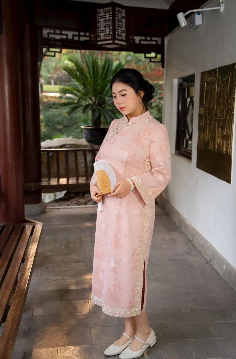 Woman in a traditional Plus Size Long Sleeve Pink Floral Qipao with Pearl Trim  dress standing in an outdoor setting with plants and a bench.