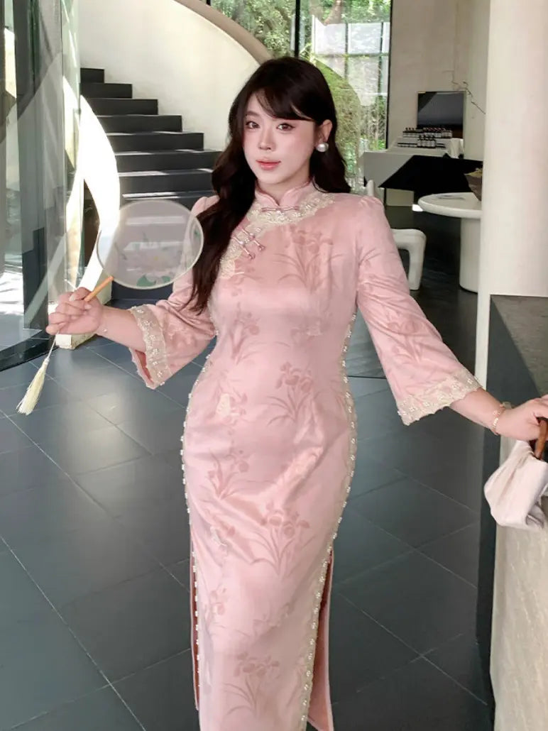 A woman wearing a light pink embroidered long sleeve plus size cheongsam/qipao dress with lace trim on the collar and sleeves, holding a round fan in an indoor modern setting.