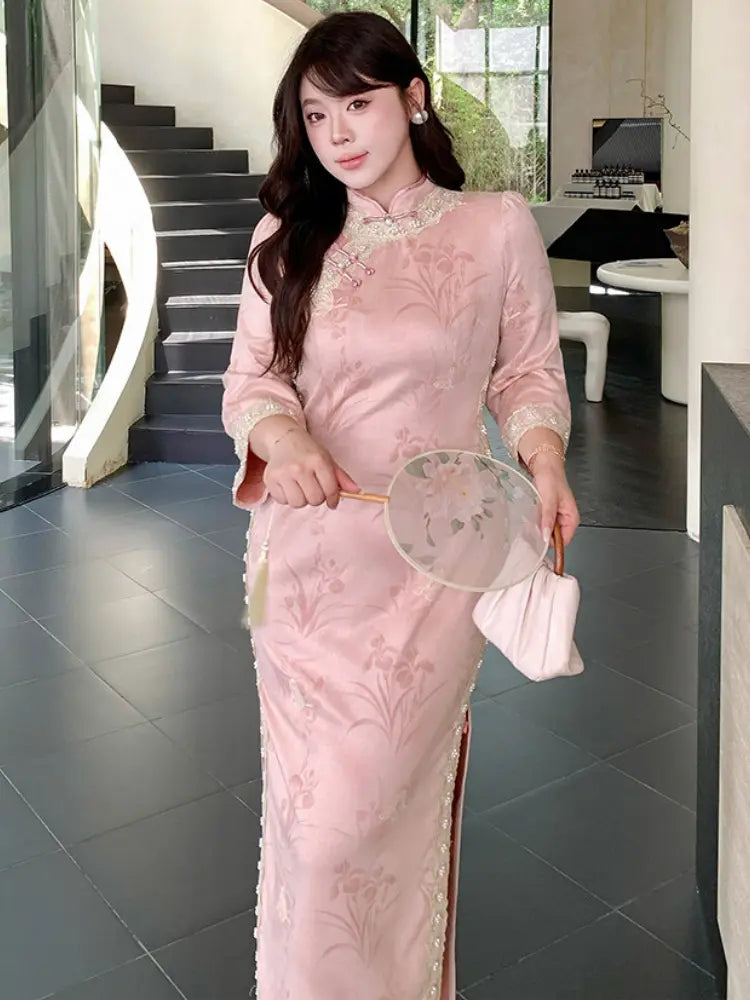 A woman wearing a light pink embroidered long sleeves cheongsam/qipao dress with floral patterns and lace trim, holding a round fan and a small matching handbag in an indoor modern setting.