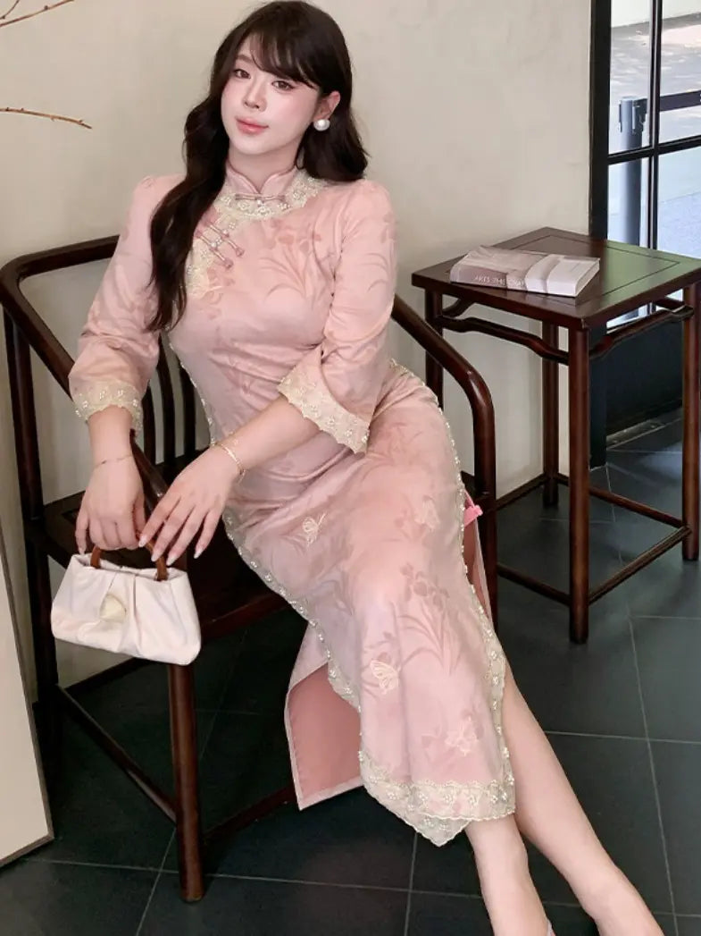 A woman wearing a light pink long sleeve embroidered cheongsam dress with lace trim, sitting on a dark wooden chair in an indoor setting.