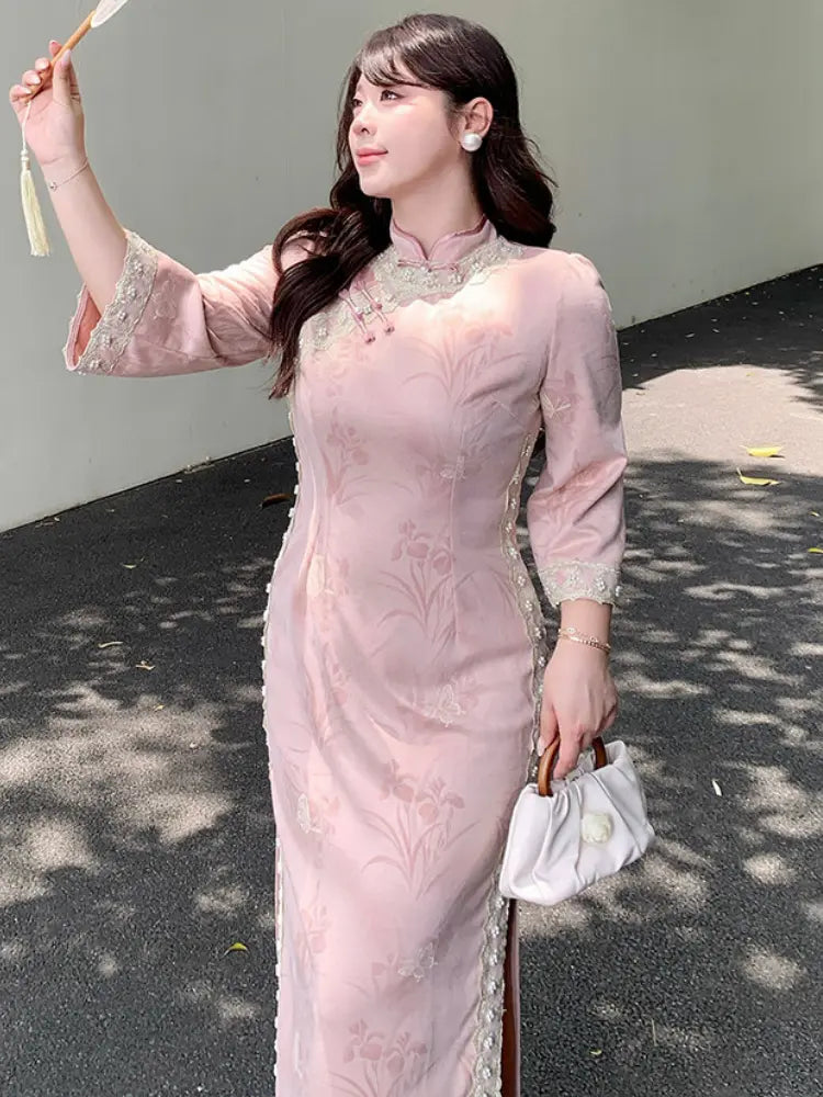 A woman wearing a light pink embroidered long sleeve plus size cheongsam/qipao dress with lace trim on the sleeves and collar, holding a small white handbag outdoors against a gray wall.