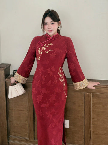 A woman wearing a red Chinese cheongsam/qipao dress with gold floral embroidery and gold-trimmed cuffs, standing indoors against a light wall and wooden cabinet.