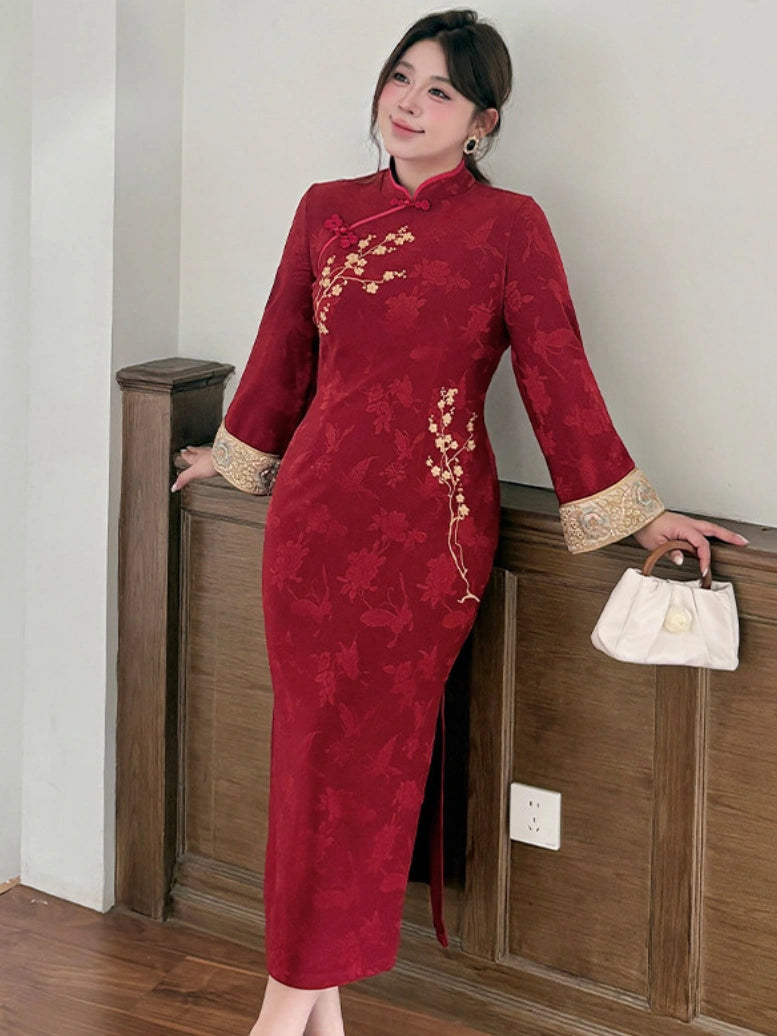A woman wearing a long-sleeved red cheongsam dress with gold floral embroidery and beige cuffs, holding a small white handbag while posing indoors.