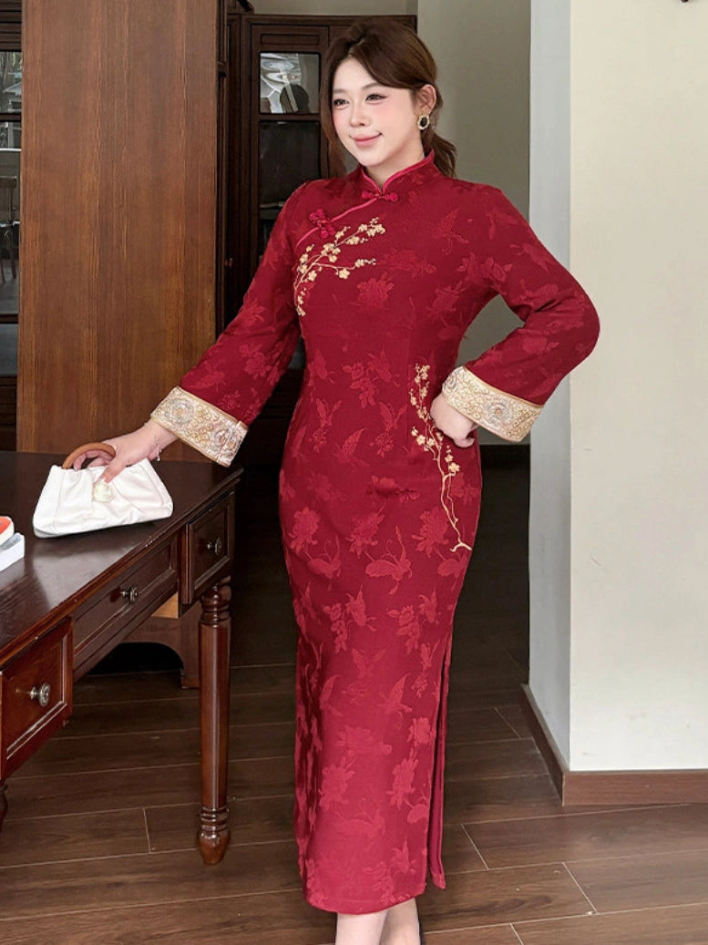 A woman wearing a long-sleeved red cheongsam dress with gold floral embroidery and beige patterned cuffs, standing in an indoor room with wooden furniture.