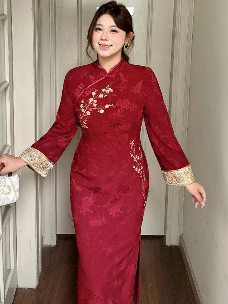 A woman wearing a long-sleeved red cheongsam dress with gold floral embroidery and gold lace cuffs, standing in a hallway.