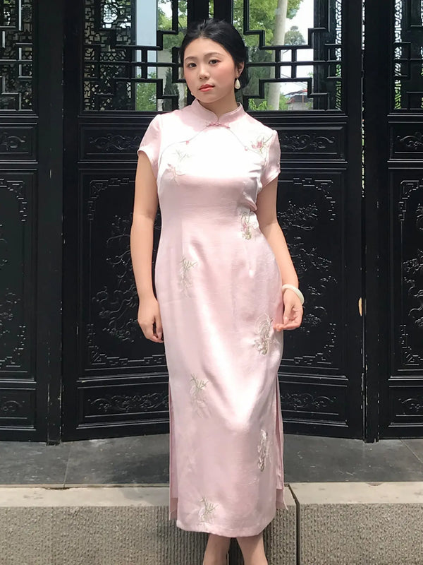A woman wearing a light pink satin cheongsam dress with floral embroidery, standing in front of a black wooden gate.