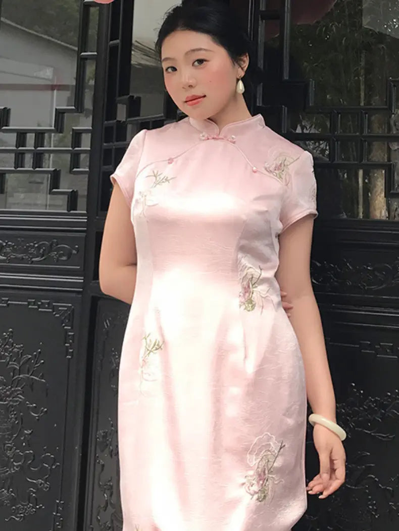 A woman wearing a light pink short-sleeve cheongsam dress with delicate floral embroidery, standing in front of a dark traditional wooden gate.