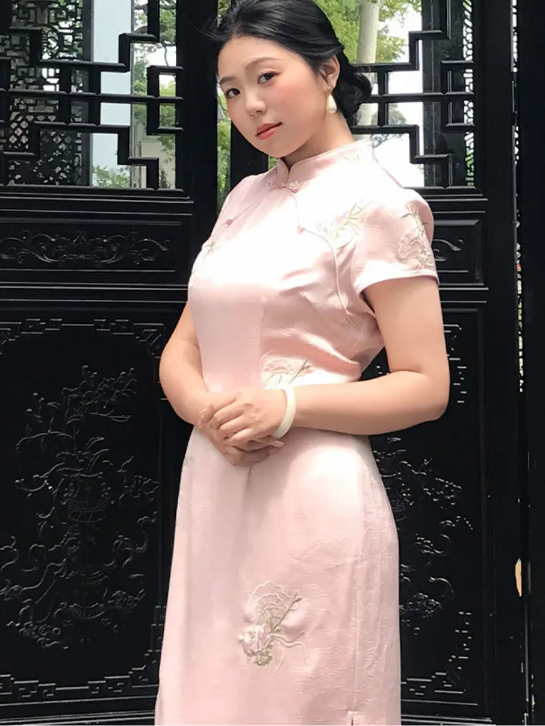 A woman wearing a light pink short-sleeved cheongsam dress with floral embroidery, standing in front of a black decorative wooden lattice screen.