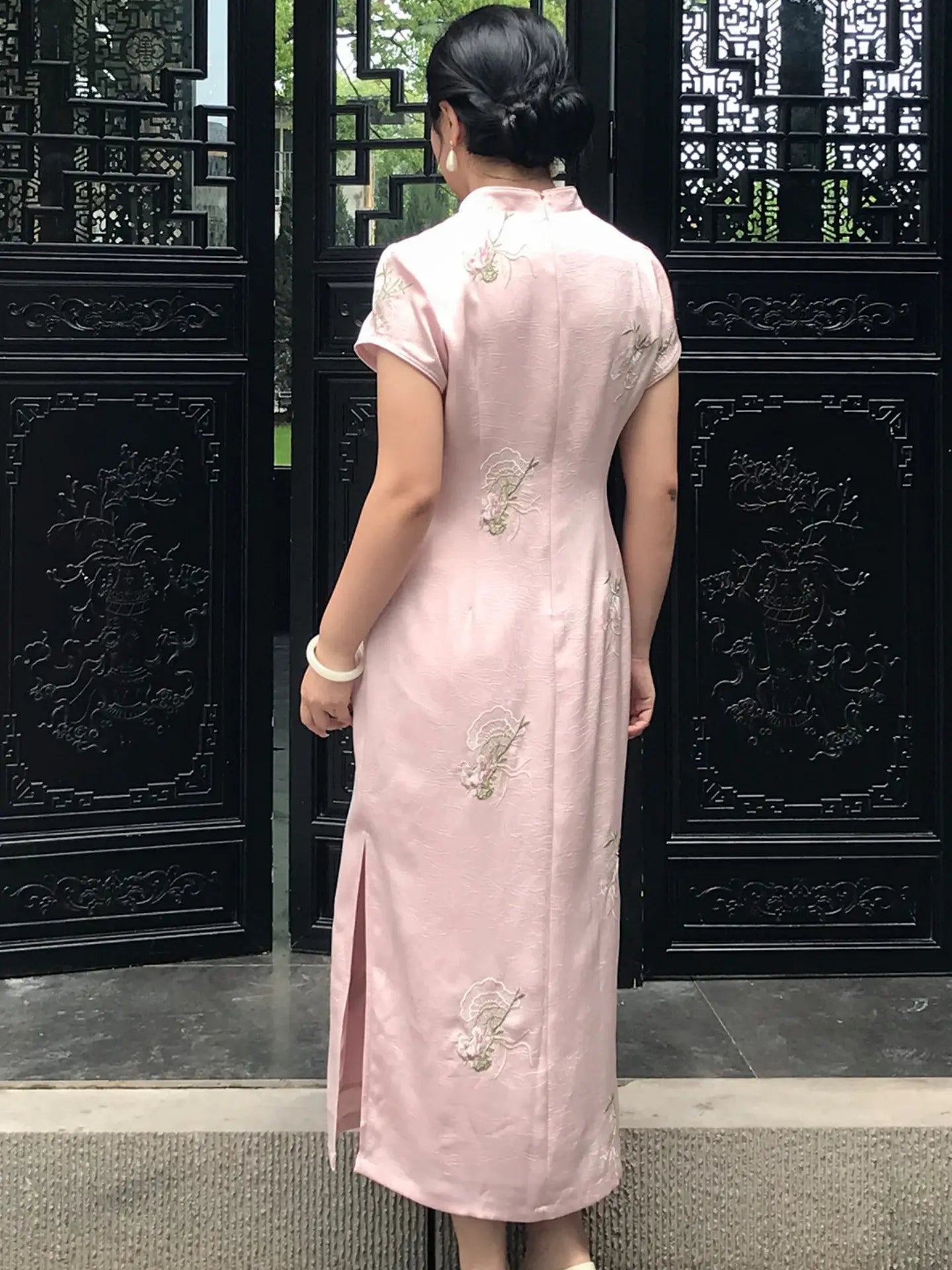 A woman viewed from behind wearing a light pink traditional Chinese cheongsam dress with silver floral embroidery, standing in front of a black ornate wooden gate.