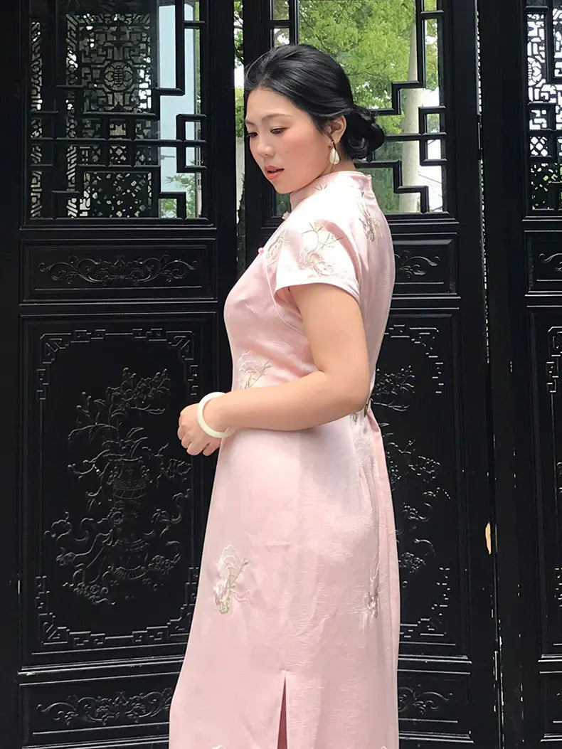 A woman wearing a light pink embroidered cheongsam dress stands in front of a black wooden lattice screen.