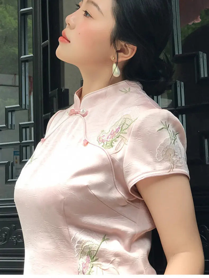 A woman wearing a light pink short-sleeve cheongsam-style dress with floral embroidery on the chest and shoulder, posed outdoors against a dark window background.
