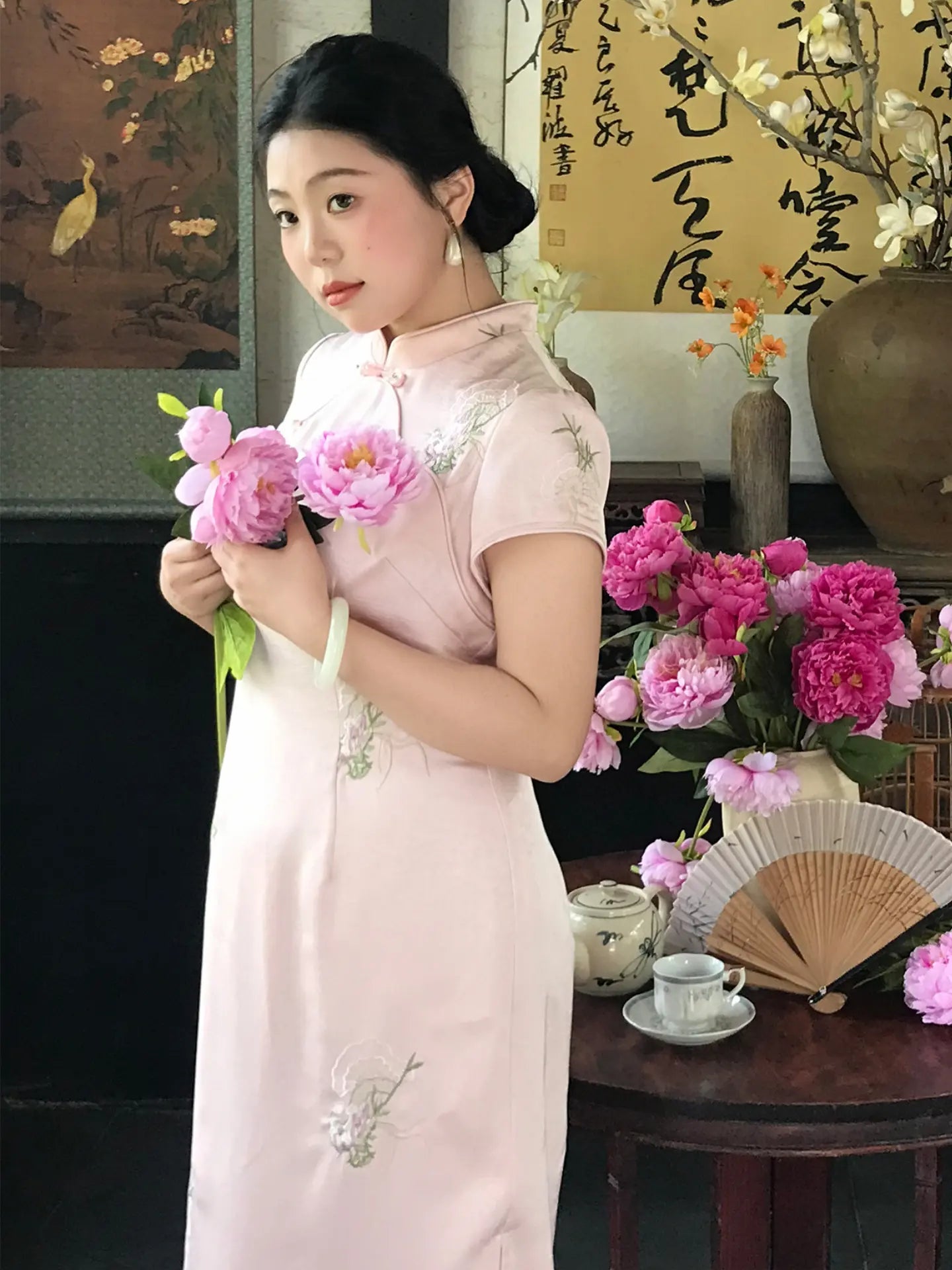 A woman wearing a pale pink traditional Chinese cheongsam dress with floral embroidery, holding pink flowers in a decorated indoor setting with a table, teacup, and calligraphy.