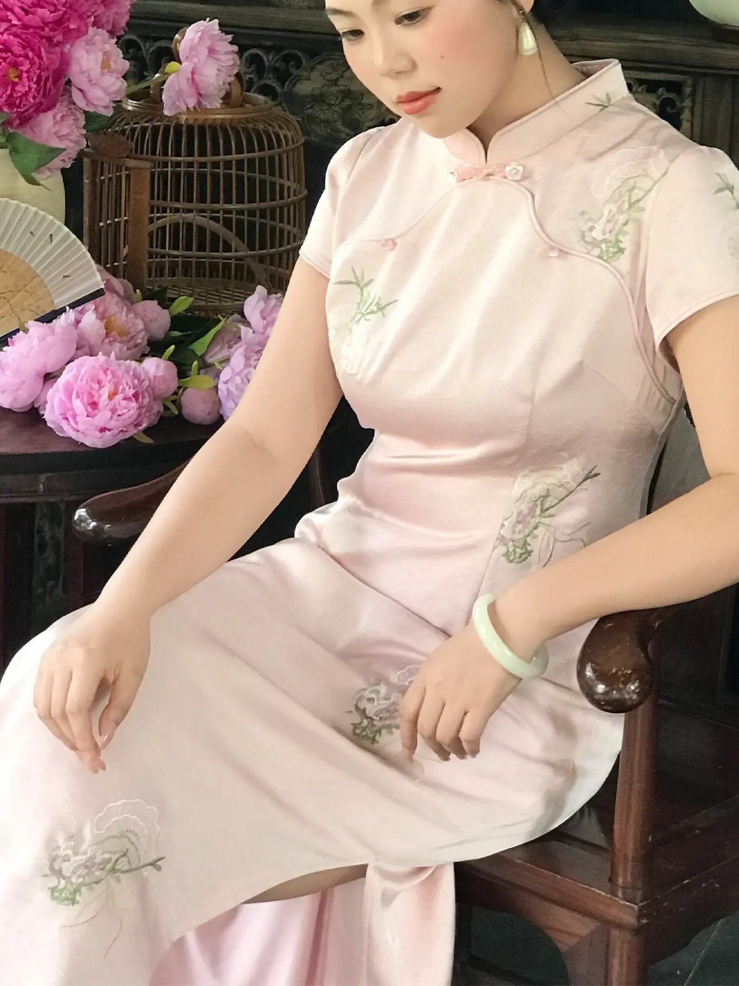 A woman wearing a light pink traditional Chinese cheongsam dress with green floral embroidery, seated in a wooden chair with pink flowers and a birdcage in the background.