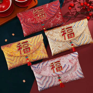 Four Chinese New Year envelope-style pouches with red tassels and embroidered Chinese characters, arranged on a dark tabletop with decorative red berries and tea cups in the background.