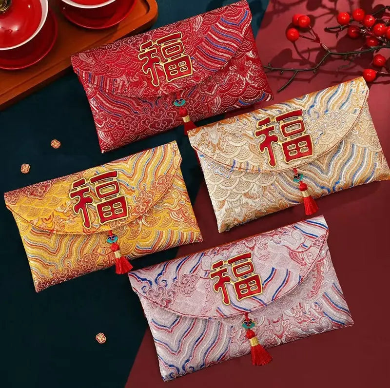 Four Chinese New Year envelope-style pouches with red tassels and embroidered Chinese characters, arranged on a dark tabletop with decorative red berries and tea cups in the background.