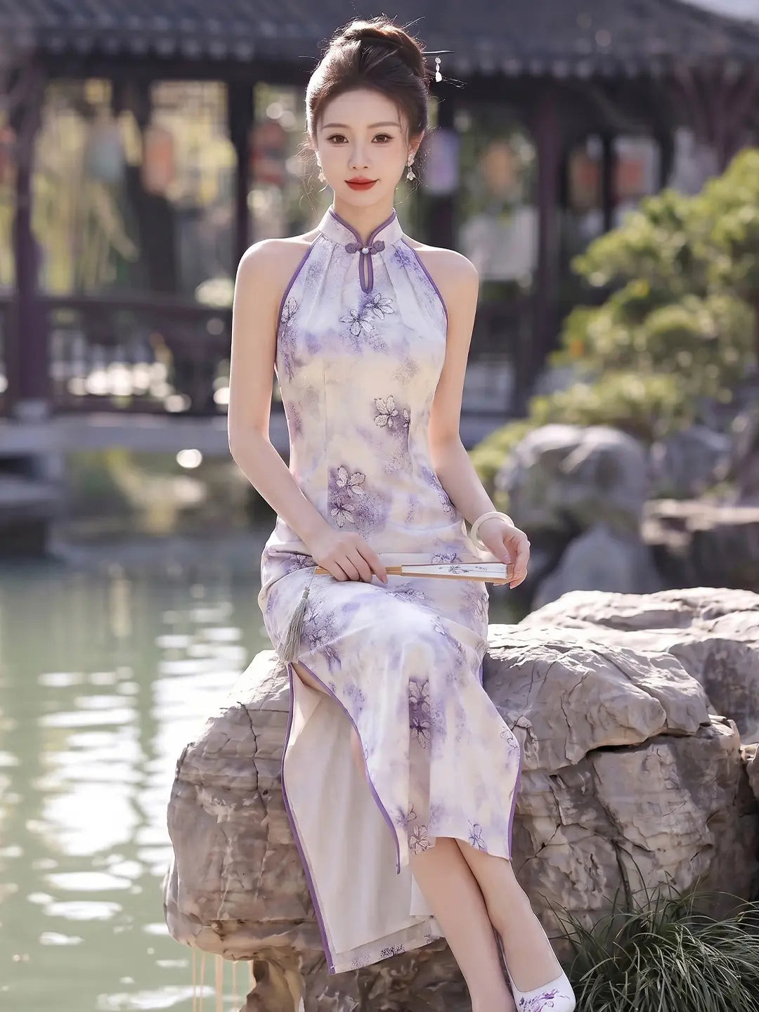 A woman wearing a sleeveless lavender and white floral print  Modern Halter Neck Jacquard Qipao cheongsam sits on a large rock by a pond in a traditional Chinese garden.