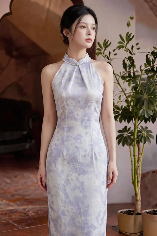 A woman wearing a sleeveless, high-neck, floor-length cheongsam-style dress with a light lavender and white watercolor pattern, standing in an indoor setting with a potted plant in the background.