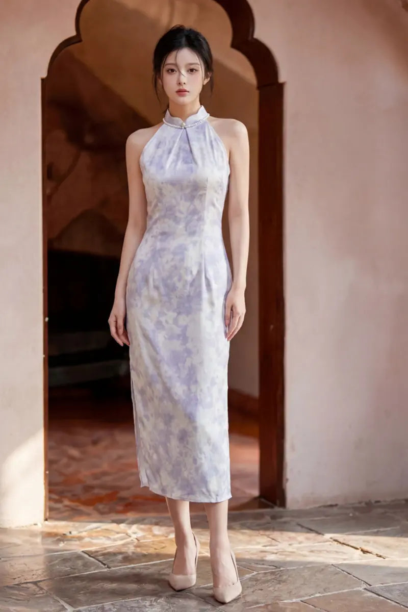 A woman stands in a doorway wearing a sleeveless, high-neck midi dress with a light lavender and white watercolor-like Modern Pearl-Embellished Halter-Style Qipao cheongsam dress. The setting features warm, earthy tones with stone flooring and arches.