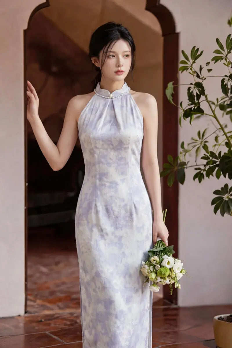 A woman wearing a sleeveless Modern Pearl-Embellished Halter-Style Qipao cheongsam dress with a light lavender and white watercolor pattern, holding a small bouquet of white and green flowers in a warm-toned indoor setting.