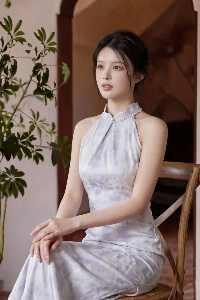 A woman wearing a sleeveless, high-neck, light lavender and white patterned Modern Pearl-Embellished Halter-Style Qipao cheongsam dress sits on a wooden chair in a warm-toned indoor setting.