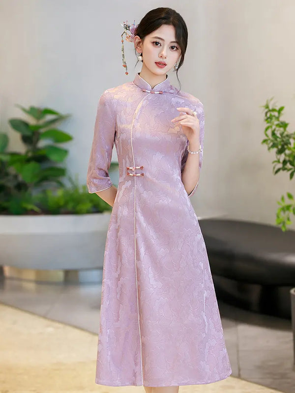 A woman wearing a lavender purple Chinese a line cheongsam dress with three-quarter sleeves and traditional button closures, standing in an indoor setting with blurred plants and furniture in the background.