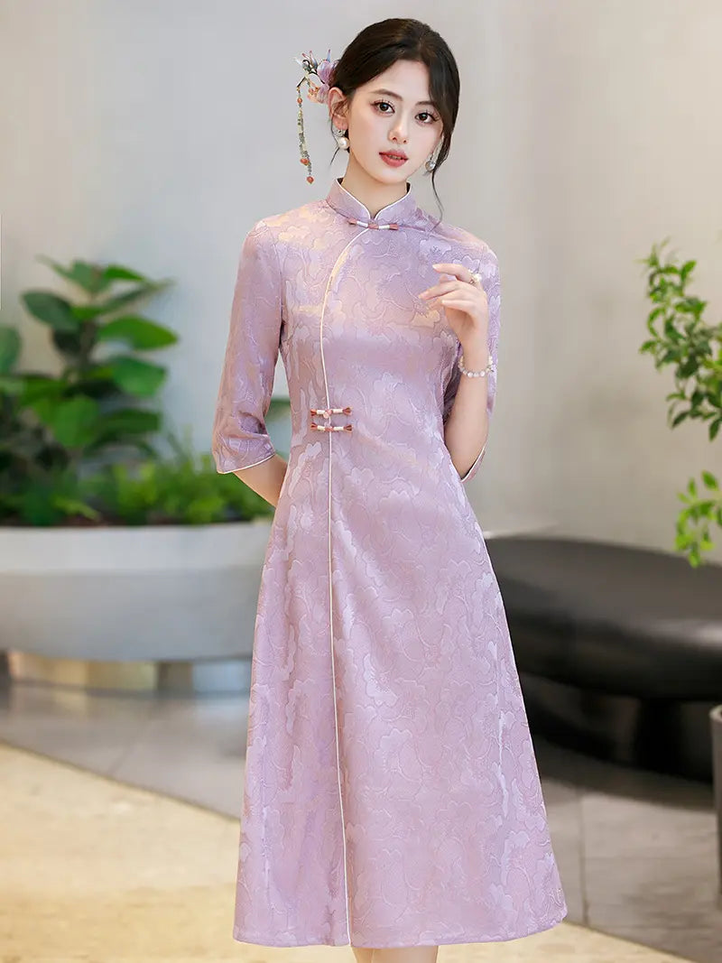 A woman wearing a lavender purple Chinese a line cheongsam dress with three-quarter sleeves and traditional button closures, standing in an indoor setting with blurred plants and furniture in the background.