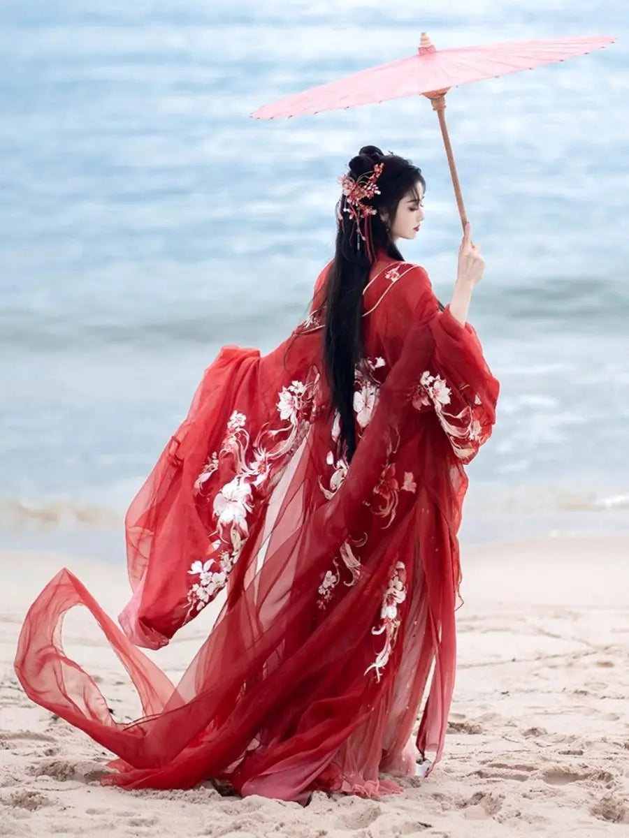 Woman in a red traditional Red Ancient Chinese Imperial Tang Dynasty Hanfu Set  with floral patterns standing on a beach holding a pink parasol.