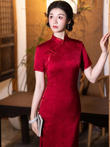A woman wearing a short-sleeved red cheongsam/qipao dress with traditional Chinese collar and frog closures, standing in a warm-toned indoor setting with wooden furniture and plants.