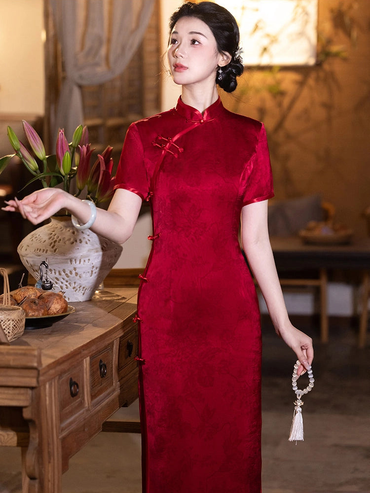 A woman wearing a short-sleeved red Bridal cheongsam/qipao dress with traditional Chinese collar and button details, standing in a warm, rustic indoor setting next to a wooden table with flowers.