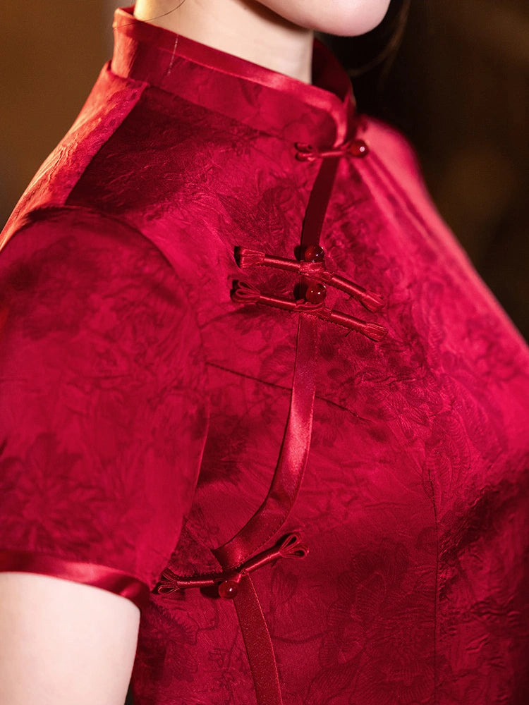 A close-up of a person wearing a short-sleeved red cheongsam-style top with a textured jacquard pattern and traditional frog closures on the front.