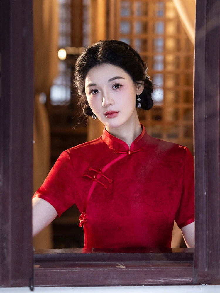 A woman wearing a red short-sleeve cheongsam-style dress with a mandarin collar and front tie, posed behind a dark wooden window frame in a warm-toned indoor setting.