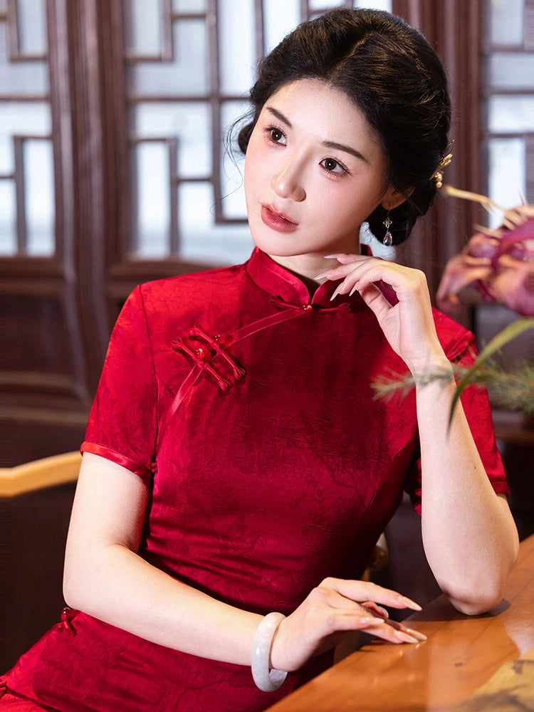 A woman wearing a red short-sleeve cheongsam-style dress with a high collar and front button placket, posed indoors against a dark wooden lattice window background.