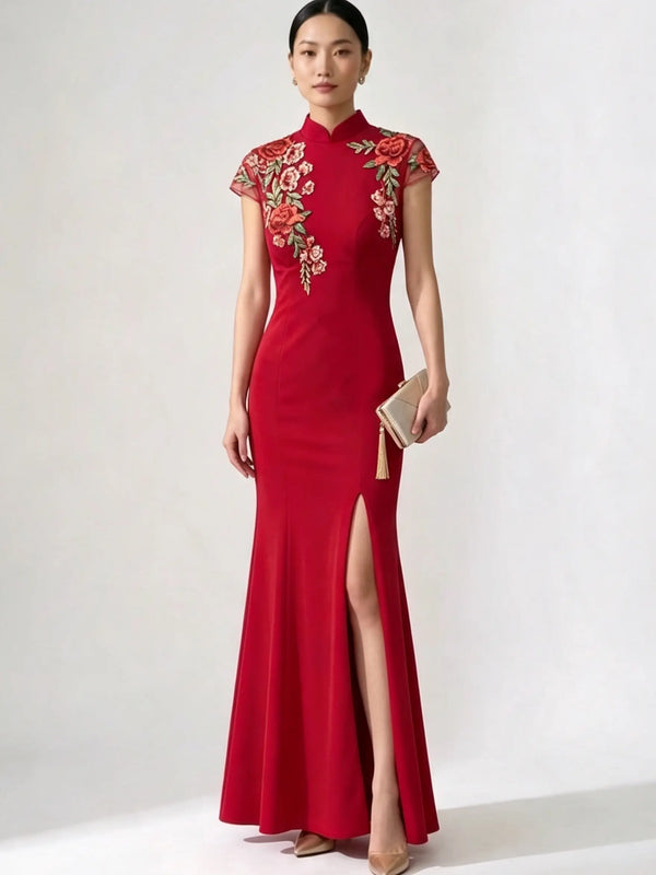 A woman models a long red cheongsam-style gown with floral embroidery on the bodice and cap sleeves, a high side slit, and a small beige clutch, photographed against a plain light gray studio background.