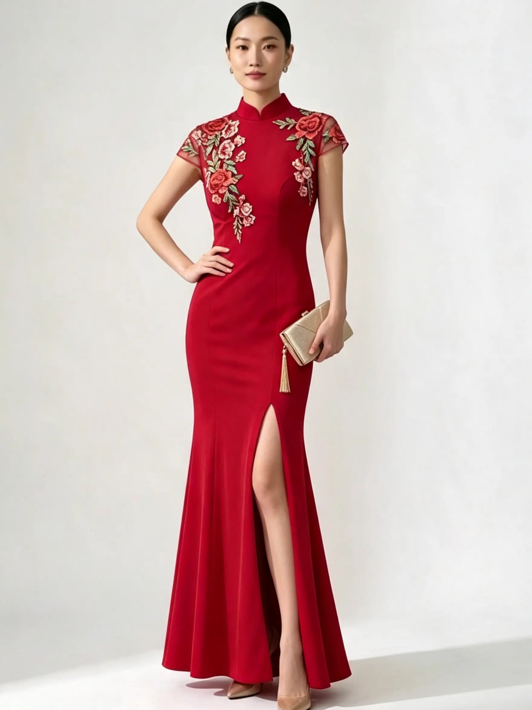 A woman models a red floor-length cheongsam-style gown with floral embroidery on the bodice and sleeves, standing against a plain light gray studio background and holding a small beige clutch.