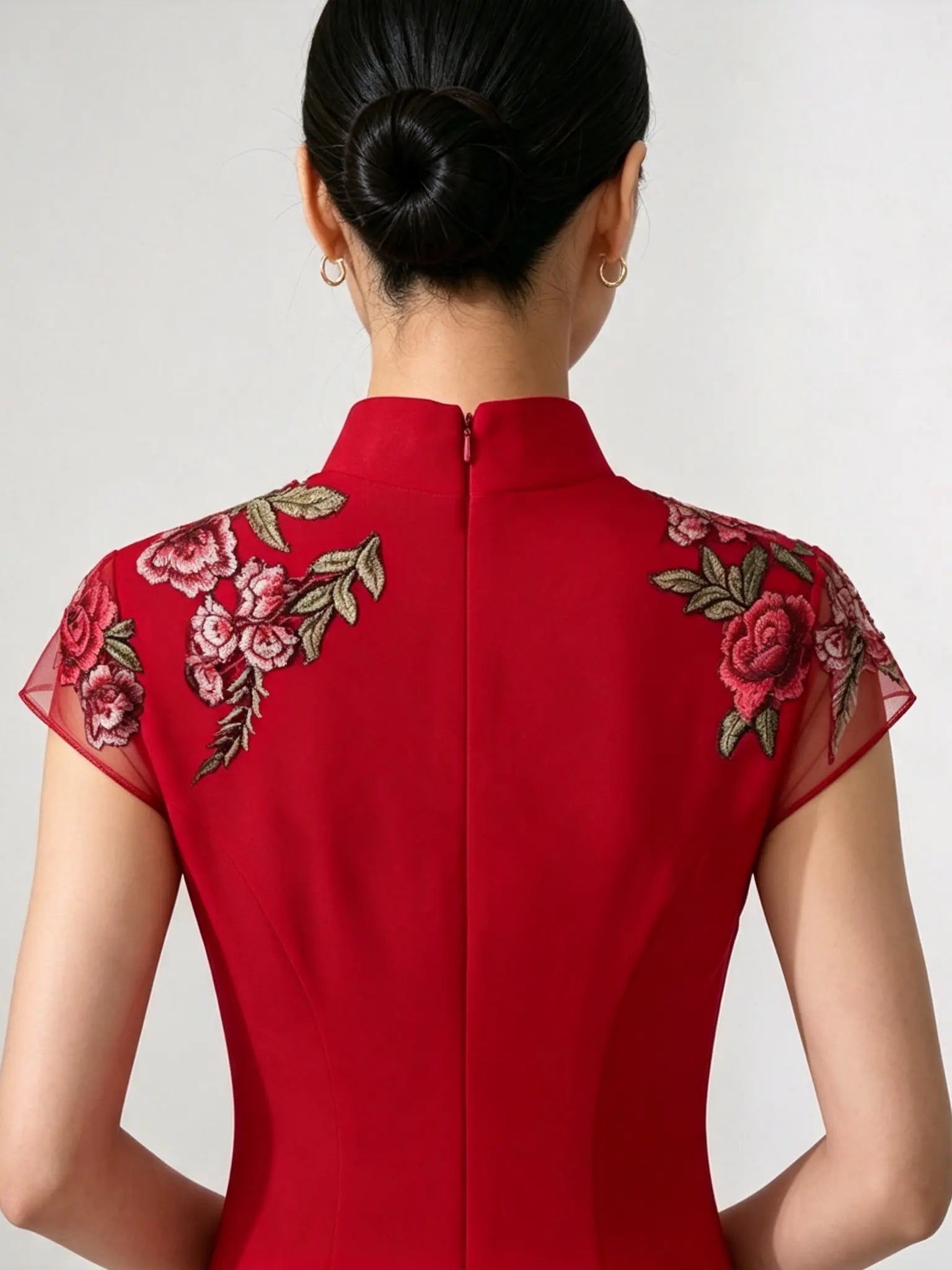 A woman viewed from the back wearing a bright red high-neck dress with floral embroidery on the shoulders. The dress has sheer cap sleeves and a central zipper.