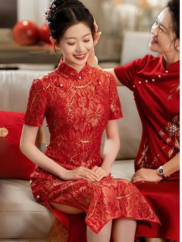 Two women wearing matching red and gold lace cheongsam/qipao dresses with gold floral detailing, seated indoors on a sofa. One woman is adjusting the other's hair, and both are smiling.