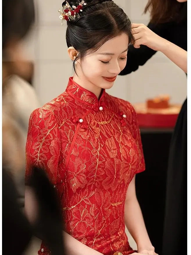 A woman wearing a red and gold lace traditional Chinese wedding cheongsam/qipao dress with gold floral detailing and pearl-like buttons, seated indoors while another person adjusts her hair.
