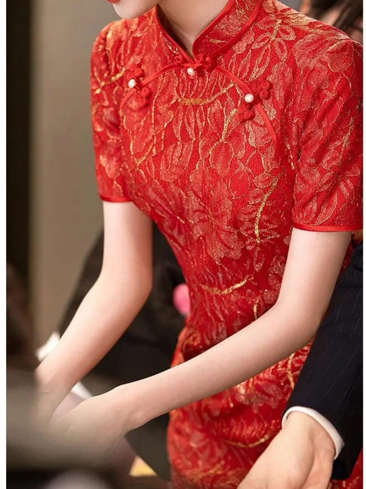 A close-up of a person wearing a red and gold short-sleeve lace cheongsam/qipao dress with gold floral embroidery and red decorative buttons at the collar.