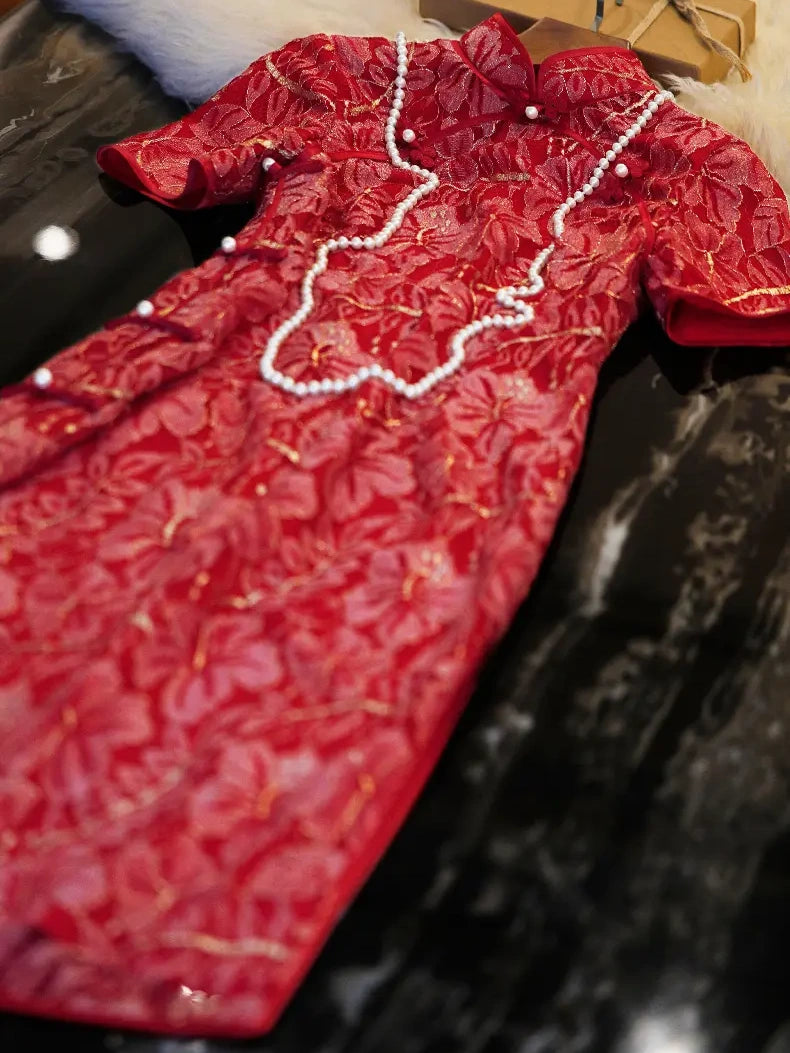 A red lace cheongsam-style dress with gold floral detailing and white pearl bead trim, laid flat on a dark reflective surface.