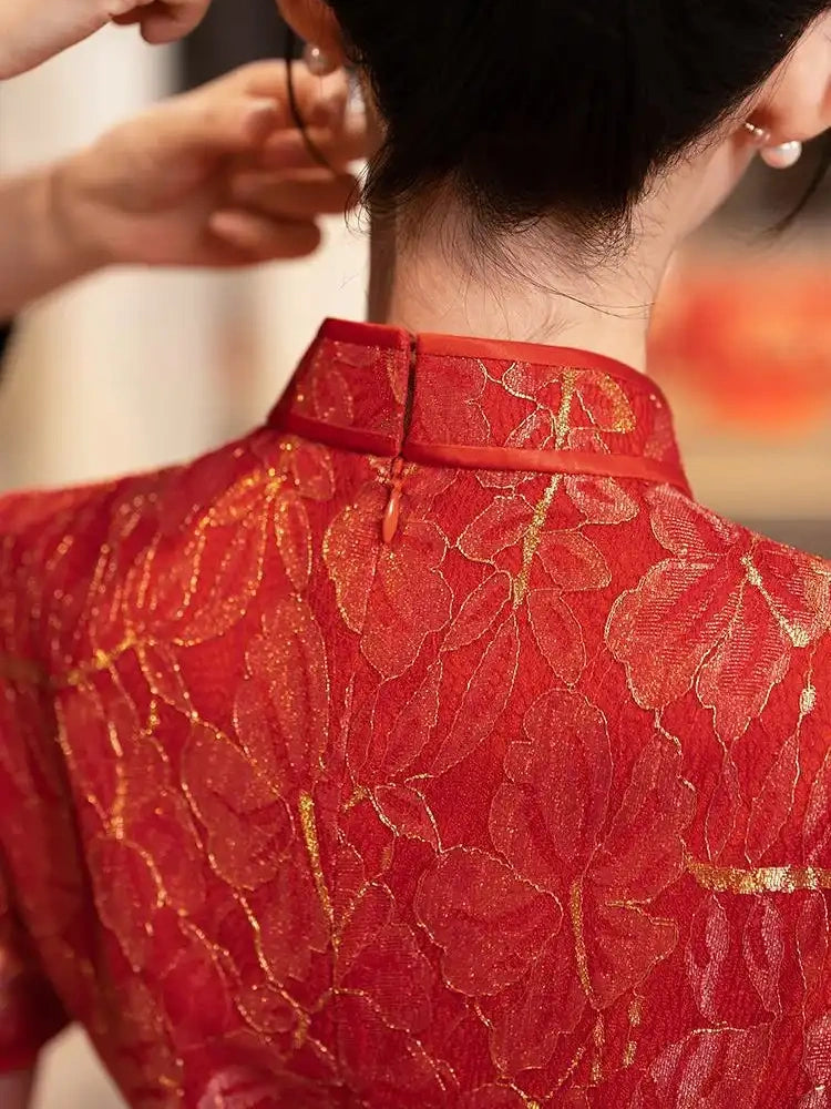Close-up back view of a person wearing a red  and gold lace  traditional chinese wedding qipao with gold floral detailing and a high collar, while another person adjusts the hair.
