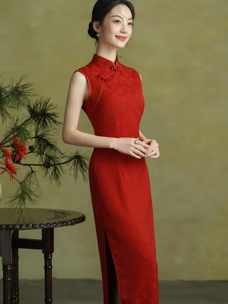 A woman wearing a sleeveless red cheongsam dress with a high slit, standing in front of a textured olive-green background next to a small table with greenery.