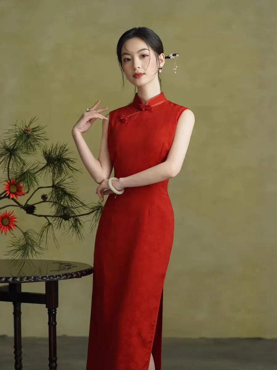 A woman wearing a sleeveless red cheongsam/qipao dress with a high side slit, standing in front of a textured olive-green background next to a small table with a pine branch and red flowers.