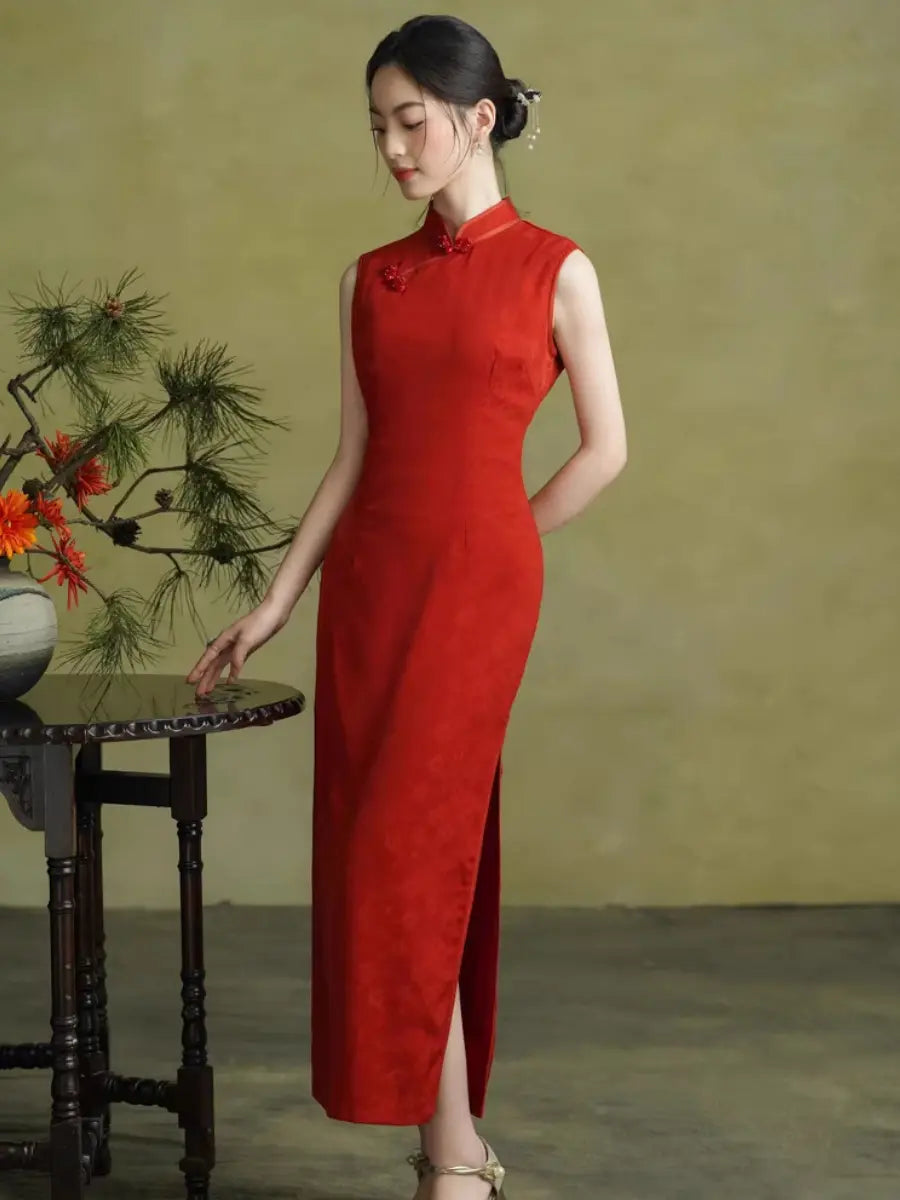 A woman wearing a sleeveless red cheongsam dress with a high side slit, standing beside a dark wooden table with a floral arrangement against a textured olive-green studio backdrop.