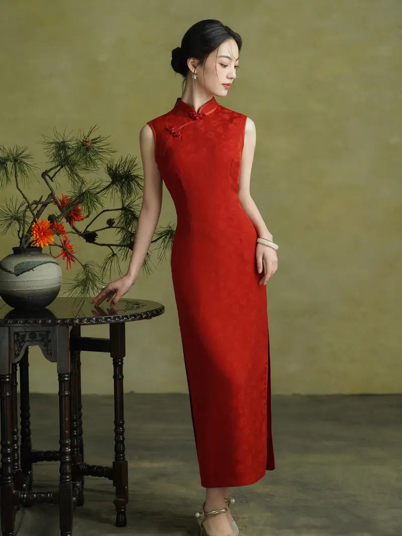 A woman wearing a sleeveless red cheongsam dress stands beside a small dark wooden table with a vase of flowers, against a muted olive-green studio backdrop.
