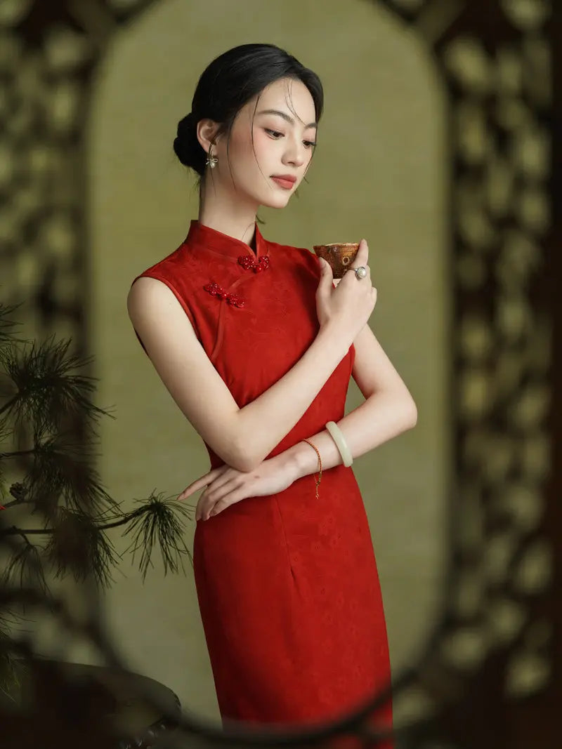 A woman wearing a sleeveless red cheongsam dress with traditional collar and button details, holding a small cup. She is posed in front of a decorative wooden screen with a soft olive-green background.