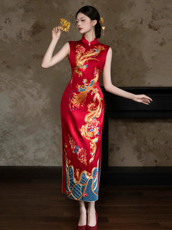 A woman models a sleeveless red wedding cheongsam/qipao dress with intricate gold, blue phoenix embroidery. She is standing in a room with a textured brown wall and dark wooden floor.