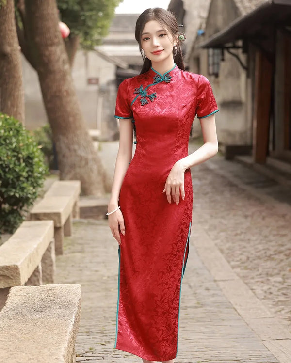 Woman in a red traditional  Short Sleeves Floral Jacquard Long Qipao Dress With Blue Trim standing on a stone path with trees and buildings in the background.