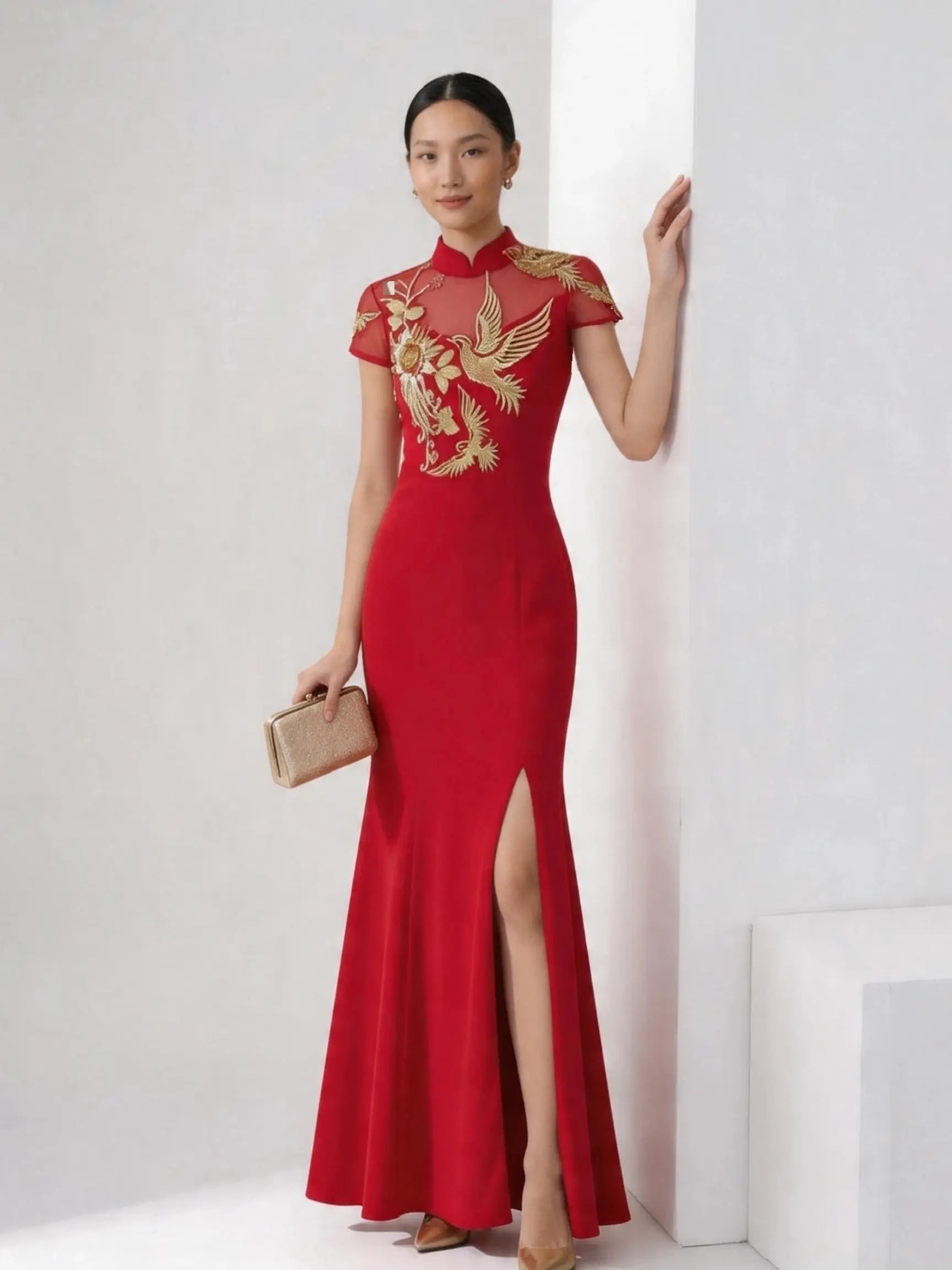 A woman models a red floor-length cheongsam-style gown with gold phoenix embroidery on the bodice and sleeves, standing in a minimalist white studio setting and holding a small beige clutch.