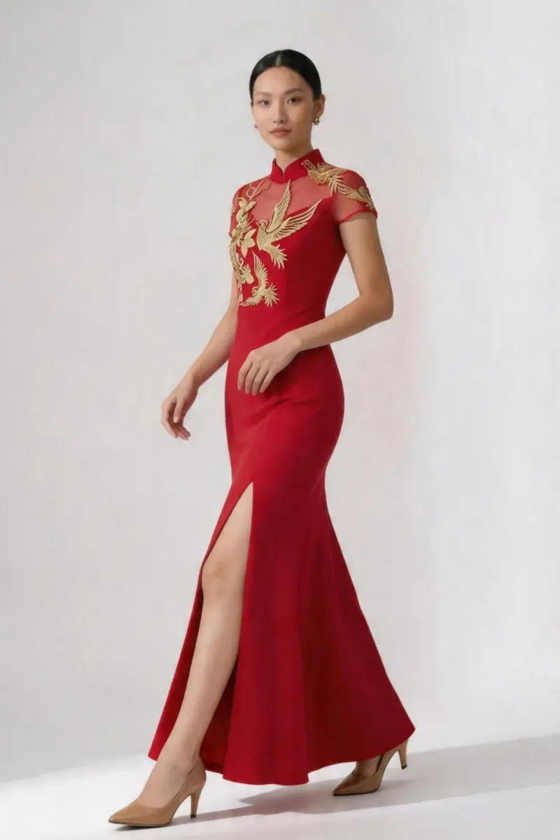 A woman modeling a red floor-length cheongsam-style gown with gold embroidered phoenix motifs on the bodice and upper skirt, photographed against a plain light gray studio background.